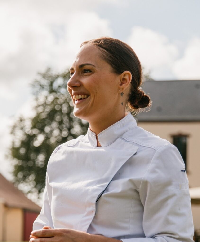 Femme chef de cuisine souriante, en veste de cuisine. Photo prise en extérieur.