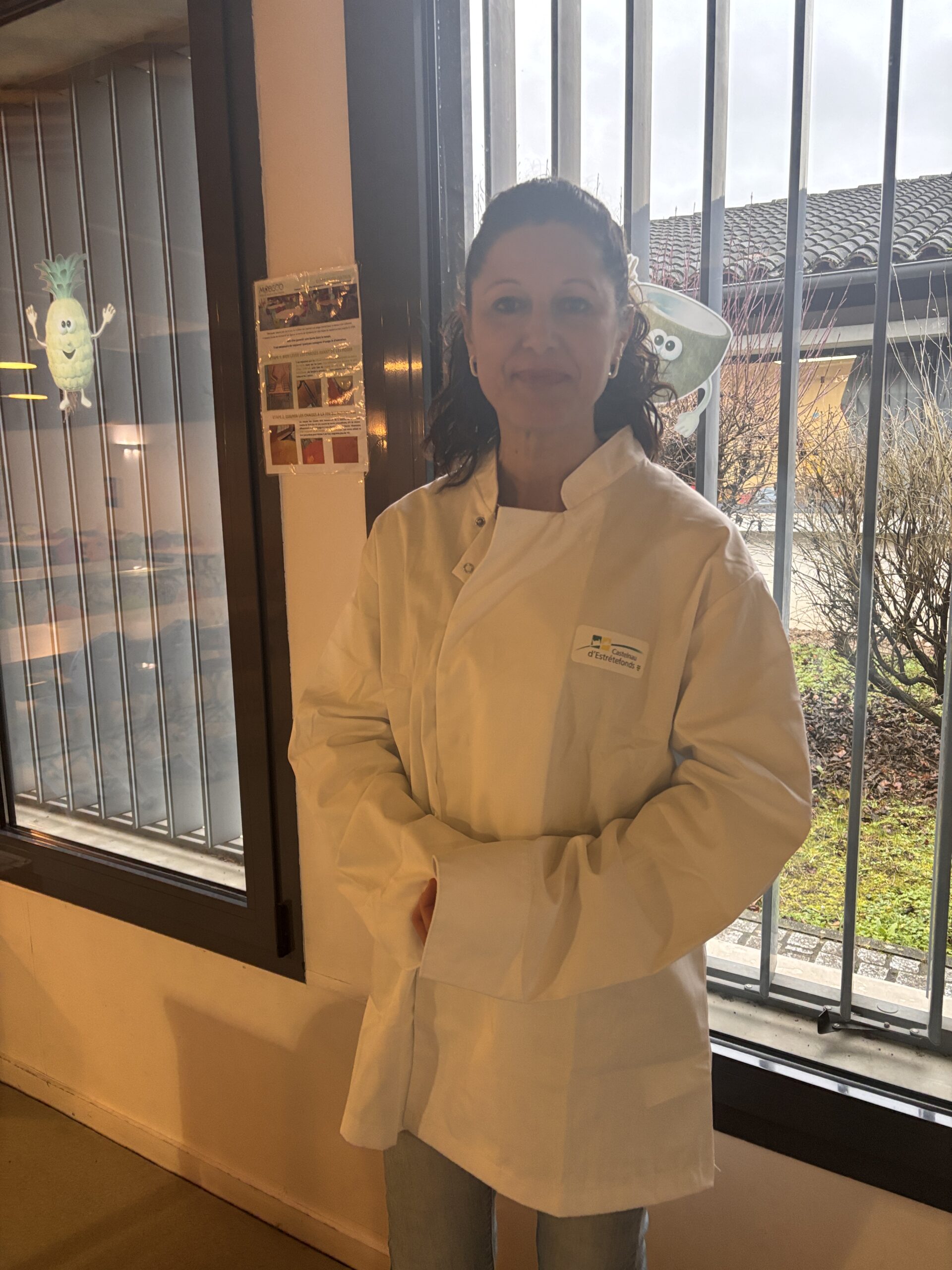 Portrait en pied, dos à une fenêtre, d'une femme agente de restauration en tenue de travail.